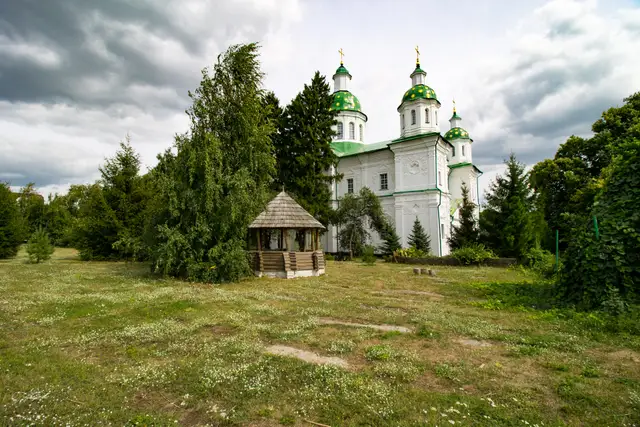 Спасо-Преображенський собор Мгарського монастиря із зеленими банями серед дерев і дерев'яна альтанка над криницею